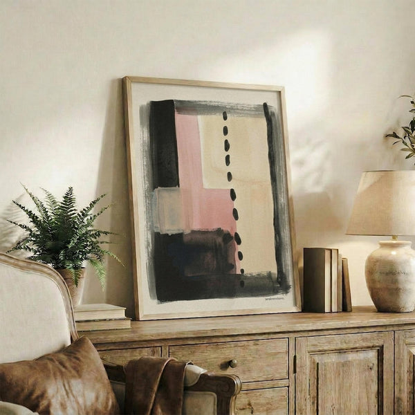 May include: Abstract art print in light wood frame displayed on rustic wooden sideboard in modern living room. Artwork features geometric blocks in pink, beige, black and gray tones with vertical dotted line detail. Scene includes fern plant, stacked books, ceramic vase, and cream upholstered chair with brown leather throw blanket.