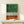 May include: Framed abstract forest artwork with green watercolor wash at top, black vertical lines resembling tree trunks on white background, and orange-brown watercolor base. Light wood frame displayed on white wooden dresser beside small wrapped gift with evergreen sprig. Minimal modern home decor styling against cream wall.