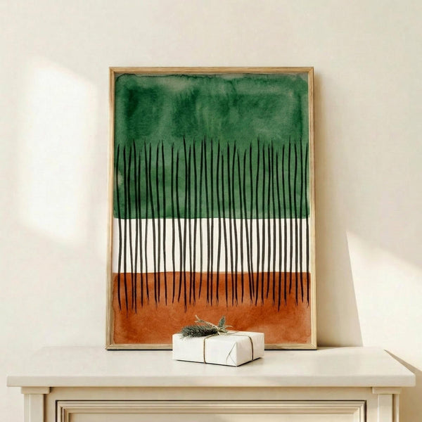 May include: Framed abstract forest artwork with green watercolor wash at top, black vertical lines resembling tree trunks on white background, and orange-brown watercolor base. Light wood frame displayed on white wooden dresser beside small wrapped gift with evergreen sprig. Minimal modern home decor styling against cream wall.