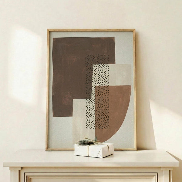 May include: Abstract geometric art print in natural wood frame leaning against white wall on light wooden surface. Artwork features overlapping brown, cream, and dotted pattern shapes in earthy tones. Small wrapped gift with evergreen sprig sits beside frame. Minimalist Scandinavian-style room setting with soft natural lighting and neutral color palette.