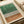 Abstract minimalist artwork featuring vertical black lines of varying heights against a watercolor background with green wash in the upper portion and warm brown tones below. The piece is displayed as a print with white border matting, lying flat on a neutral surface alongside rolled paper tubes, showing the artwork's clean geometric design and organic color transitions.
