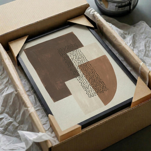 May include: Abstract geometric art print in earth tones featuring overlapping curved shapes in brown and tan with dotted stippled texture pattern. Black frame with white mat sitting in cardboard shipping box with wooden dowels and protective tissue paper wrapping.