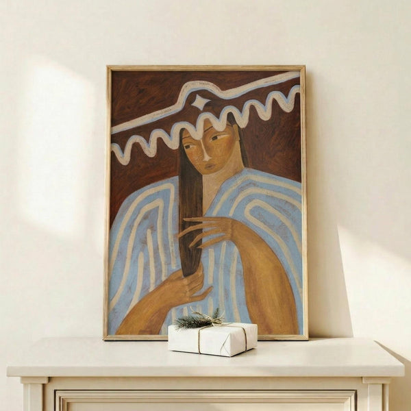 May include: Framed artwork displayed on light wood shelf showing portrait of woman with long dark hair in blue and white striped garment against brown background with white wavy design elements. Small white wrapped gift with greenery sits beside the natural wood frame. Clean white wall background.