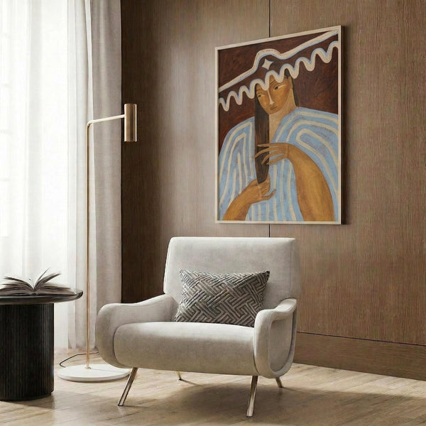 May include: Framed portrait artwork featuring a person in blue and white striped clothing against a dark reddish-brown background with flowing white forms above. The painting has warm earth tones and appears to be in a modernist style. Shown hanging on wood-paneled wall above a cream-colored upholstered armchair with geometric patterned pillow, next to a brass floor lamp and dark round side table with open book.