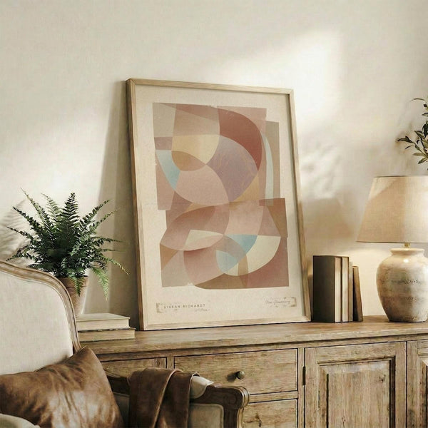 May include: Abstract geometric art print in muted earth tones featuring overlapping circular and angular shapes in soft browns, peaches, and dusty blue, displayed in light wood frame on rustic wooden sideboard alongside fern plant, ceramic vase, and leather chair in warm-lit living room setting.