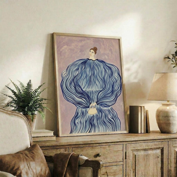 May include: Framed artwork featuring a woman with brown hair in a bun wearing an elaborate blue flowing dress or gown with wave-like patterns against a soft purple background. The piece is displayed on a rustic wooden sideboard alongside books, a ceramic vase, and a fern plant, with a beige upholstered chair visible in the foreground of this neutral-toned living space.