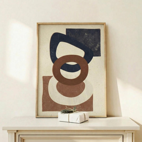 May include: Framed abstract geometric art print featuring overlapping organic shapes in navy blue, terracotta brown, and cream colors on textured beige paper, displayed on light wood console table with small wrapped gift box beside it against neutral wall