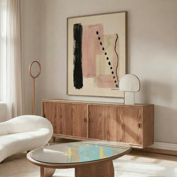 May include: Abstract art print with pink, black, and cream brushstrokes displayed above a walnut sideboard in a minimalist living room. The framed artwork features geometric shapes, dotted lines, and soft color blocks. Modern white curved chair and glass coffee table with blue accents complete the neutral-toned interior setting with natural lighting from nearby window.