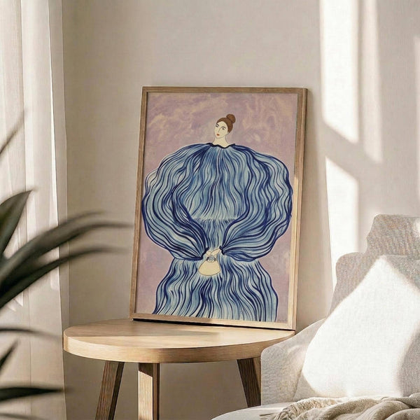 May include: Framed art print displayed on round wooden side table in bright living room with beige chair and plant. Artwork shows woman with elaborate blue flowing dress or fabric draped around her against soft purple background. Natural wood frame catches sunlight streaming through window onto neutral walls.
