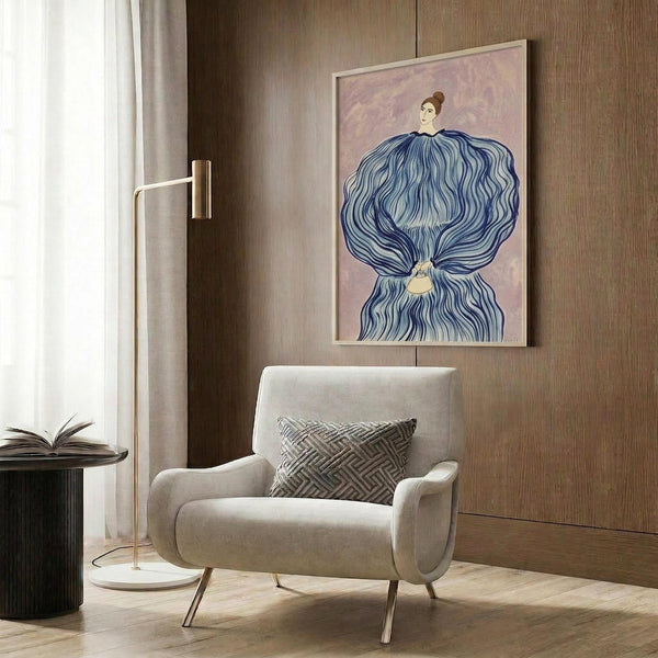May include: Framed art print featuring a woman in an elaborate blue flowing gown with dramatic fabric draping, displayed above a beige upholstered armchair with geometric pillow in a modern room setting with wood paneled walls, gold floor lamp, and dark round side table with open book.