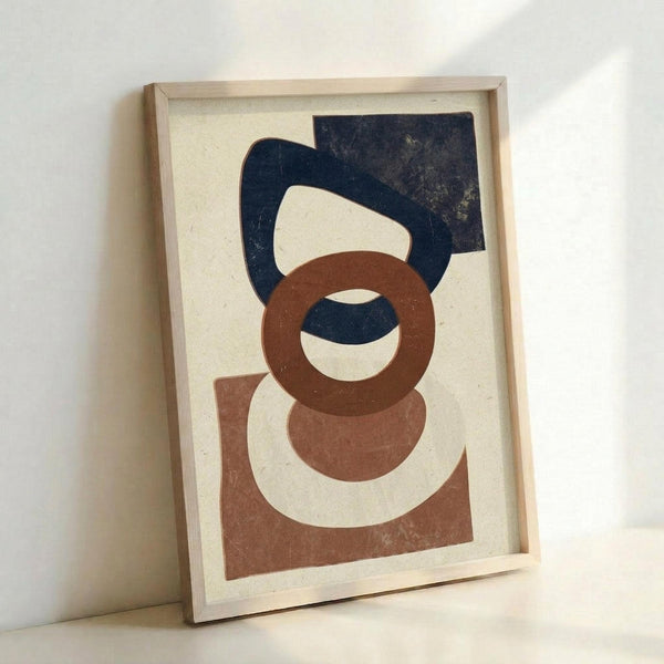 May include: Framed abstract geometric artwork leaning against white wall. Features overlapping organic shapes in navy blue, terracotta brown, and cream on textured beige background. Natural wood frame with visible grain. Soft natural lighting creates subtle shadows on wall and frame surface.