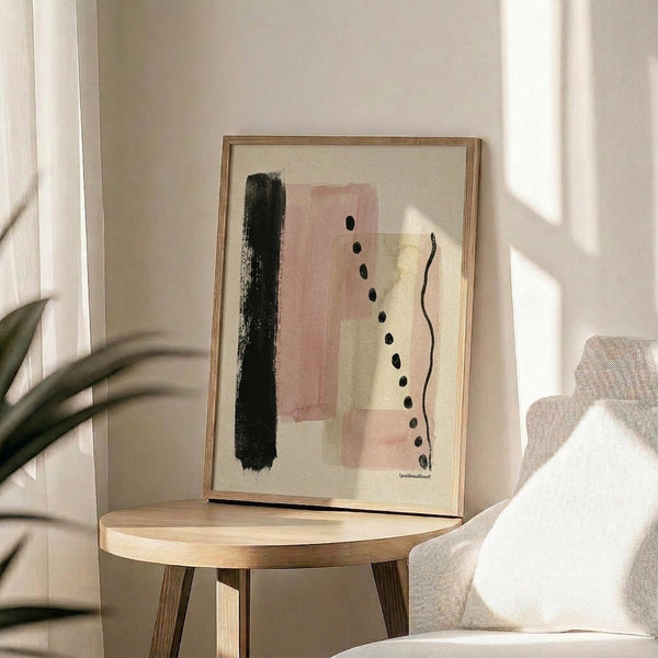 May include: Modern abstract art print with pink rectangles, black brushstroke, dotted line, and wavy line on cream background, displayed in light wood frame on round wooden side table next to beige upholstered chair. Natural sunlight streams across white walls creating geometric shadows, with green plant leaves visible in corner of minimalist living room setting.