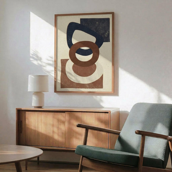 May include: Abstract geometric artwork with overlapping navy blue and terracotta brown shapes on cream background, displayed in thin wood frame above mid-century modern walnut credenza. Sage green upholstered armchair with wood arms sits nearby, white ceramic table lamp on credenza, natural sunlight casting shadows across white wall in contemporary living room setting.