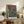 May include: Framed poster displayed on rustic wooden sideboard in cozy living room setting. Artwork features anime-style illustration of soldier character standing on rain-soaked Neo-Tokyo street with mecha robot towering behind, rendered in teal and orange tones with Japanese text. Natural wood frame complements weathered furniture, fern plant, ceramic vase, and cream armchair visible in warm, softly-lit interior space.