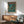 May include: Framed poster displayed above wooden sideboard in modern living room setting. Artwork features anime-style illustration with soldier character and large mech robot on rainy futuristic city street. Poster has Japanese text at top and teal, orange color scheme. Room shows mid-century furniture including sage green armchair with wooden arms, round coffee table, and white ceramic lamp. Natural sunlight streams across white walls.