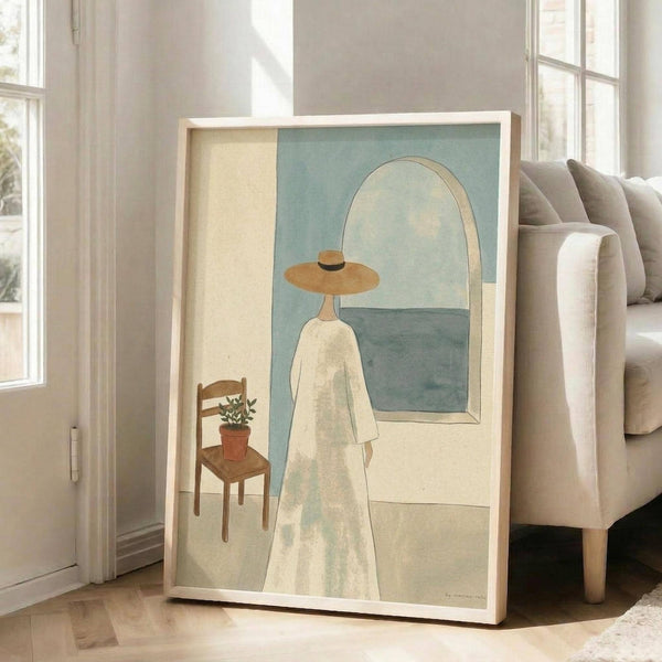 May include: Large framed poster print leaning against white wall next to beige sofa. Artwork shows figure in white dress and wide-brimmed hat viewed from behind, looking through arched window toward blue-green seascape. Small wooden chair with potted plant sits nearby. Soft, muted color palette with cream, sage green, and earth tones. Minimalist illustration style in light wood frame.