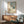 May include: Mid-century modern living room with wooden credenza, sage green upholstered armchair, and white table lamp. Framed artwork shows woman in wide-brimmed hat and flowing white dress standing by window overlooking blue sea, with wooden chair and potted plant nearby. Warm sunlight casts geometric shadows across white wall.