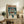 May include: Framed anime poster featuring a young character holding an umbrella in the rain with a large mecha robot looming behind them, displayed on a rustic wooden sideboard alongside potted fern, ceramic vase, and stacked books in a cozy living room setting with beige armchair