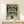 May include: Framed anime poster displayed on light wood shelf against white wall, featuring a young character holding an umbrella in the rain with a large mecha robot looming behind them. Japanese text at top in orange lettering. Small white wrapped gift box sits in front of the natural wood frame. Artwork shows detailed illustration in blues, oranges, and earth tones with dramatic lighting and rain effects.
