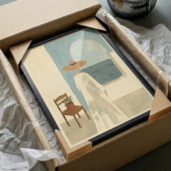May include: Framed art print in black frame showing minimalist interior scene with figure in wide-brimmed hat looking through arched window toward ocean, wooden chair with small potted plant, rendered in soft beige and sage green tones. Print packaged in cardboard box with protective wrapping, surrounded by shipping materials on white fabric surface.