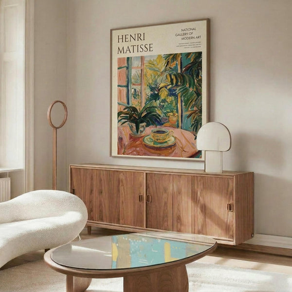 May include: Henri Matisse exhibition poster displayed above wooden sideboard in modern living room. Poster shows colorful still life with teacup, plants, and window in Matisse's distinctive style with vibrant greens, yellows, and coral tones. Room features white curved chair, oval glass coffee table, brass floor lamp, and neutral walls with natural lighting.
