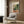 May include: Henri Matisse exhibition poster featuring colorful fauve-style interior scene with teacup, saucer, plants and window, displayed in light wooden frame on warm brown wood-paneled wall above beige upholstered armchair with geometric patterned throw pillow, alongside brass floor lamp and dark side table with open book
