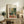 May include: Framed Henri Matisse exhibition poster displayed on rustic wooden sideboard in cozy living room setting. The artwork shows vibrant indoor scene with plants, teacup, and window in characteristic Fauvist style with bold greens, oranges, and blues. Natural wood frame complements warm interior with fern plant, ceramic vase, books, and brown leather furniture visible.