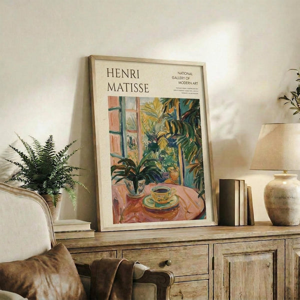 May include: Framed Henri Matisse exhibition poster displayed on rustic wooden sideboard in cozy living room setting. The artwork shows vibrant indoor scene with plants, teacup, and window in characteristic Fauvist style with bold greens, oranges, and blues. Natural wood frame complements warm interior with fern plant, ceramic vase, books, and brown leather furniture visible.