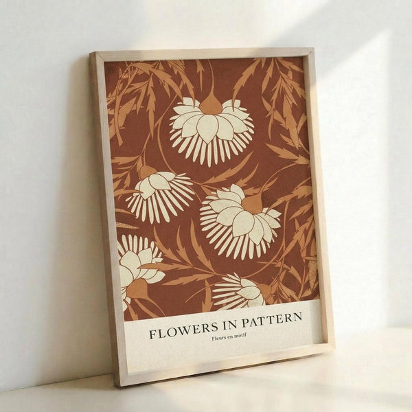 May include: A framed botanical art print leaning against a white wall, featuring stylized cream-colored flowers with radiating petals on a warm brown background with golden foliage patterns. The print has "FLOWERS IN PATTERN" and "Fleurs en motif" text at the bottom on a cream border, displayed in a natural wood frame with visible grain texture.