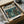 May include: Vintage-style Japanese poster featuring a military pilot in olive green uniform standing in a flooded, neon-lit cityscape. The artwork displays muted teal and orange tones with a retro aesthetic. The framed print sits in an opened cardboard box with white tissue paper padding on a neutral surface.
