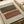 Abstract geometric artwork featuring layered horizontal bands in earthy tones of rust orange, olive green, and cream with a dotted halftone pattern running through the center. The composition sits on textured gray paper with visible fiber specks, displayed alongside rolled white paper tubes on a light wooden surface.