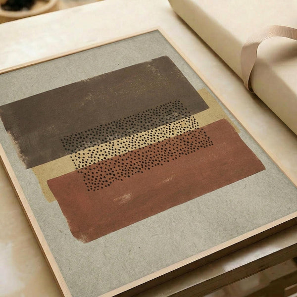 Abstract geometric artwork featuring layered horizontal bands in earthy tones of rust orange, olive green, and cream with a dotted halftone pattern running through the center. The composition sits on textured gray paper with visible fiber specks, displayed alongside rolled white paper tubes on a light wooden surface.