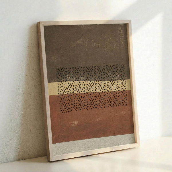 May include: Framed abstract art print leaning against white wall, featuring horizontal bands in brown, tan, rust orange, and cream with scattered black dots creating a stippled pattern across the center. Light wood frame, textured painted surfaces with subtle shimmer, positioned on light surface casting soft shadow.