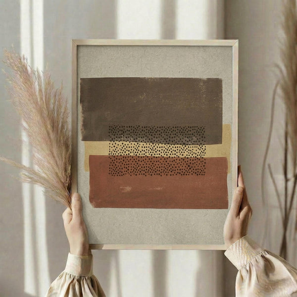 May include: Hands holding a framed abstract art print featuring horizontal bands in brown, yellow, and terracotta with dotted pattern details, displayed against white curtains with dried pampas grass visible to the left, showing natural lighting and neutral home decor styling.