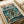 May include: Vintage-style poster with Japanese text and cyberpunk artwork displayed in white frame on cream-colored surface next to rolled paper. The poster features two armored figures in a futuristic cityscape with tall buildings, lightning, and teal-green sky. Art style resembles retro anime or manga with weathered, aged paper texture and muted color palette of browns, teals, and oranges.