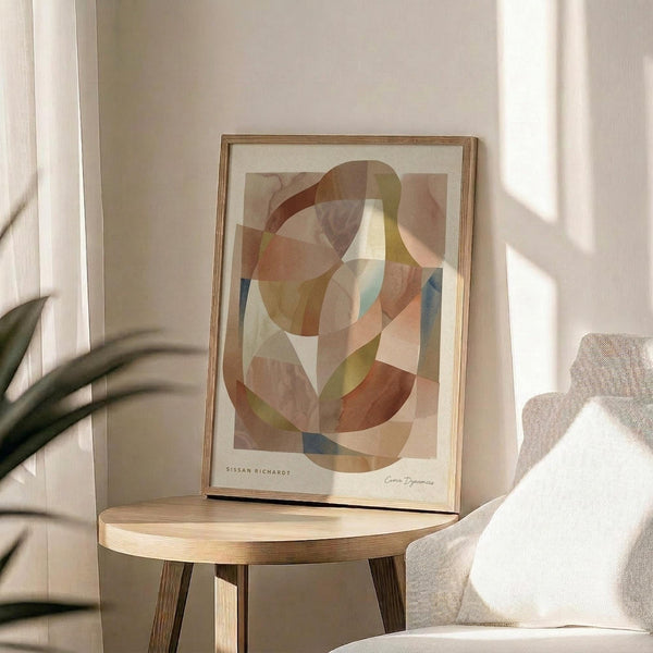 May include: Abstract art print with overlapping curved shapes in warm earth tones including terracotta, sage green, cream, and muted blue, displayed in a thin wood frame on a round wooden side table next to a beige upholstered chair, with soft natural lighting and plant shadows visible on white walls.