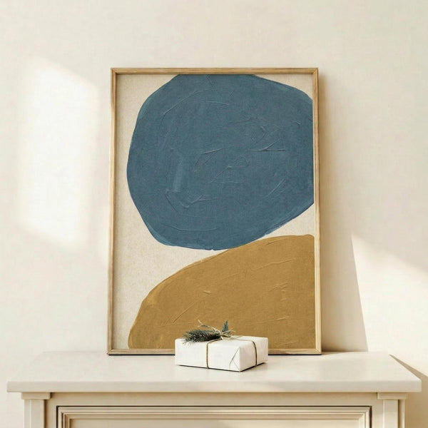 May include: Abstract artwork in wooden frame featuring large teal blue circle and golden mustard curved shape on cream background, displayed on light wood console table with small wrapped gift decorated with evergreen sprig against white wall