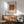 May include: Framed abstract art print with red brushstroke shape covered in green dots against cream background with vertical stripes, displayed above wooden sideboard with table lamp, next to green upholstered mid-century modern chair in sunlit room with white walls.