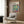May include: Henri Matisse museum poster displayed on wood-paneled wall above cream upholstered armchair with geometric pillow. Poster shows colorful open window scene with tropical plants, blue shutters, and pink table. Modern brass floor lamp and dark round side table with open book complete the styled living room setting.