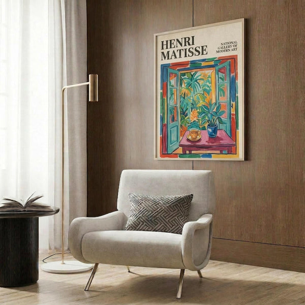 May include: Henri Matisse museum poster displayed on wood-paneled wall above cream upholstered armchair with geometric pillow. Poster shows colorful open window scene with tropical plants, blue shutters, and pink table. Modern brass floor lamp and dark round side table with open book complete the styled living room setting.