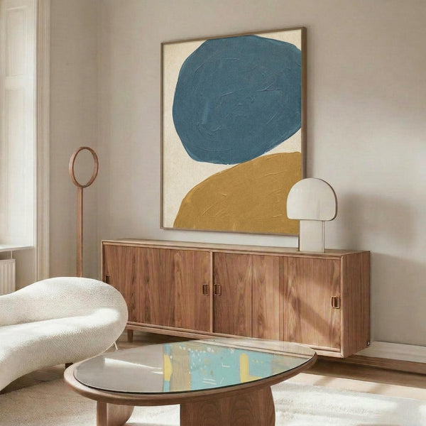 May include: Abstract poster with large teal blue circle and golden yellow curved shape on cream background, displayed in thin wooden frame above mid-century walnut credenza in modern living room with white sculptural chair, brass floor lamp, and oval glass coffee table