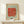 May include: Framed abstract art print featuring a red brushstroke shape with green dots on blue and white striped background, displayed on light wood console table next to small white wrapped gift with evergreen sprig, against cream wall with natural lighting