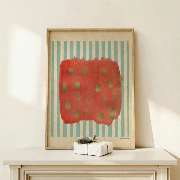 May include: Framed abstract art print featuring a red brushstroke shape with green dots on blue and white striped background, displayed on light wood console table next to small white wrapped gift with evergreen sprig, against cream wall with natural lighting