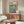 May include: Henri Matisse exhibition poster featuring colorful open window scene with tropical plants, teacup on pink table, displayed above mid-century walnut credenza in modern living room with white chair, glass coffee table, and brass floor lamp on light walls