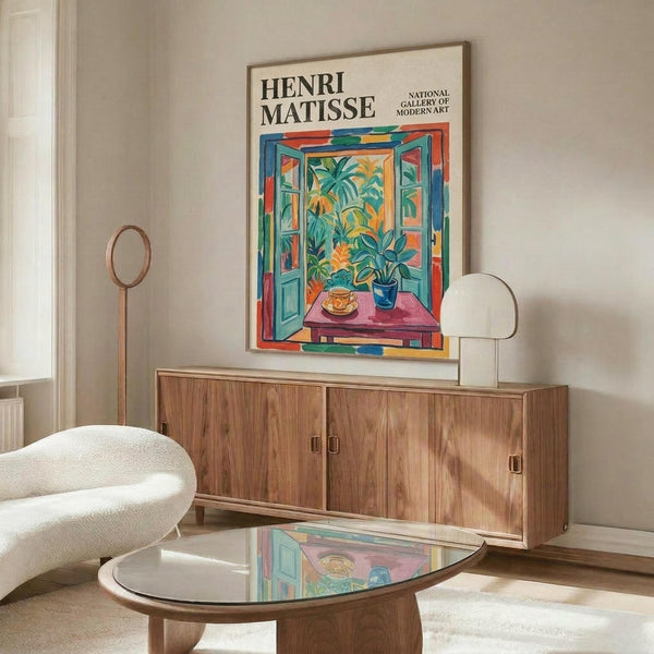May include: Henri Matisse exhibition poster featuring colorful open window scene with tropical plants, teacup on pink table, displayed above mid-century walnut credenza in modern living room with white chair, glass coffee table, and brass floor lamp on light walls
