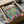 May include: Henri Matisse art print in black frame being unpacked from cardboard shipping box with protective tissue paper. The colorful poster shows tropical plants near an open window in vibrant blues, greens, oranges and pinks in Matisse's distinctive fauve style. Natural lighting on white surface with glass vessel visible in background corner.