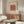May include: Abstract art print with red polka-dotted shape on light blue striped background, displayed in thin frame above mid-century modern walnut credenza. Room features cream upholstered chair, brass floor lamp, white table lamp, and oval glass coffee table on light wooden floors against neutral walls.