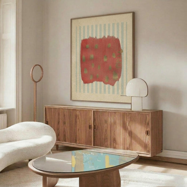 May include: Abstract art print with red polka-dotted shape on light blue striped background, displayed in thin frame above mid-century modern walnut credenza. Room features cream upholstered chair, brass floor lamp, white table lamp, and oval glass coffee table on light wooden floors against neutral walls.