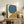 May include: Abstract art print with large teal blue circle and mustard yellow curved shape on cream background, displayed in light wood frame on rustic wooden console table alongside fern plant, books, and ceramic vases in neutral living room setting with beige armchair.