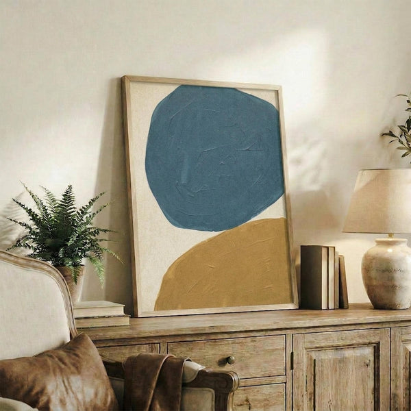 May include: Abstract art print with large teal blue circle and mustard yellow curved shape on cream background, displayed in light wood frame on rustic wooden console table alongside fern plant, books, and ceramic vases in neutral living room setting with beige armchair.